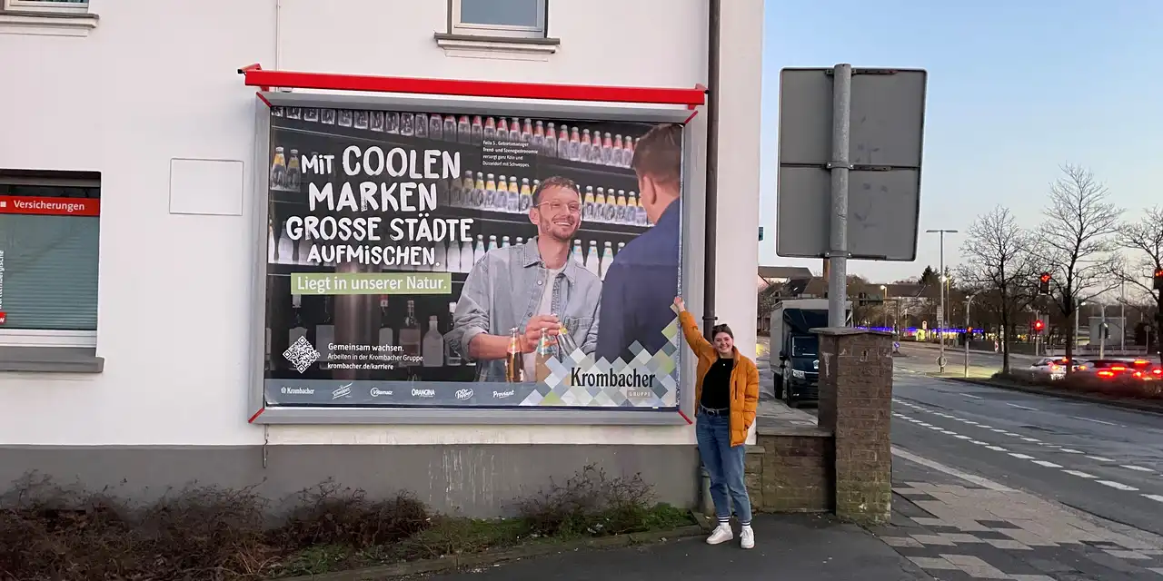 Eine Frau steht vor einem Plakat von Krombacher, das an einer Hauswand angebracht ist.