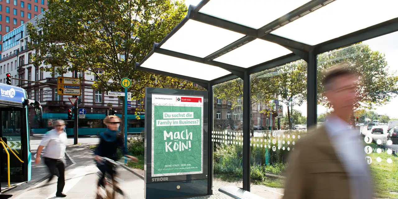 Ein Plakat mit dem Satz "Mach Köln!" an einer Bushaltestelle.