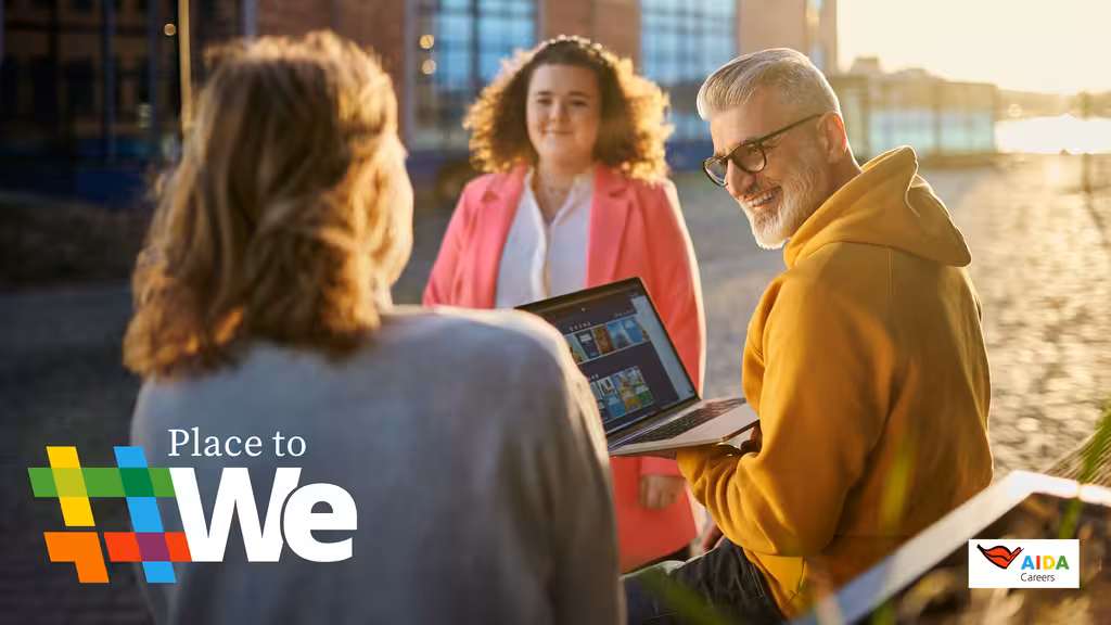 3 AIDA - Mitarbeiter sitzen nebeneinander in der Sonne und schauen gemeinsam lachend auf einen Laptop. 