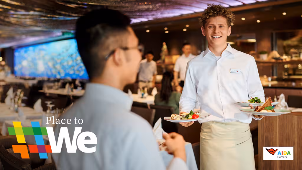 Ein AIDA Mitarbeiter im weißen Hemd und Schürze serviert lachend drei gefüllte Teller.