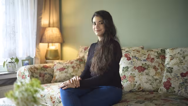 Eine junge Frau sitzt auf einem geblümten Sofa, hat die Beine übereinander geschlagen und ihre Hände darauf gelegt. Sie lacht in die Kamera.