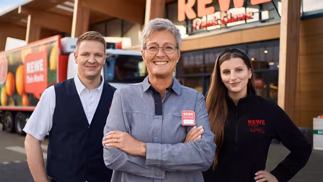 Drei Rewe-Mitarbeiter stehen nebeneinander vor einer Filiale.
