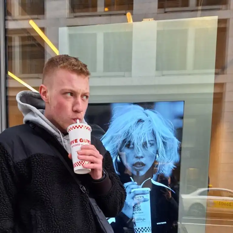 Simon Steffens, ein Mitarbeiter der Castenow GmbH, steht vor einem Schaufenster und trinkt aus einem Becher mit Strohhalm.