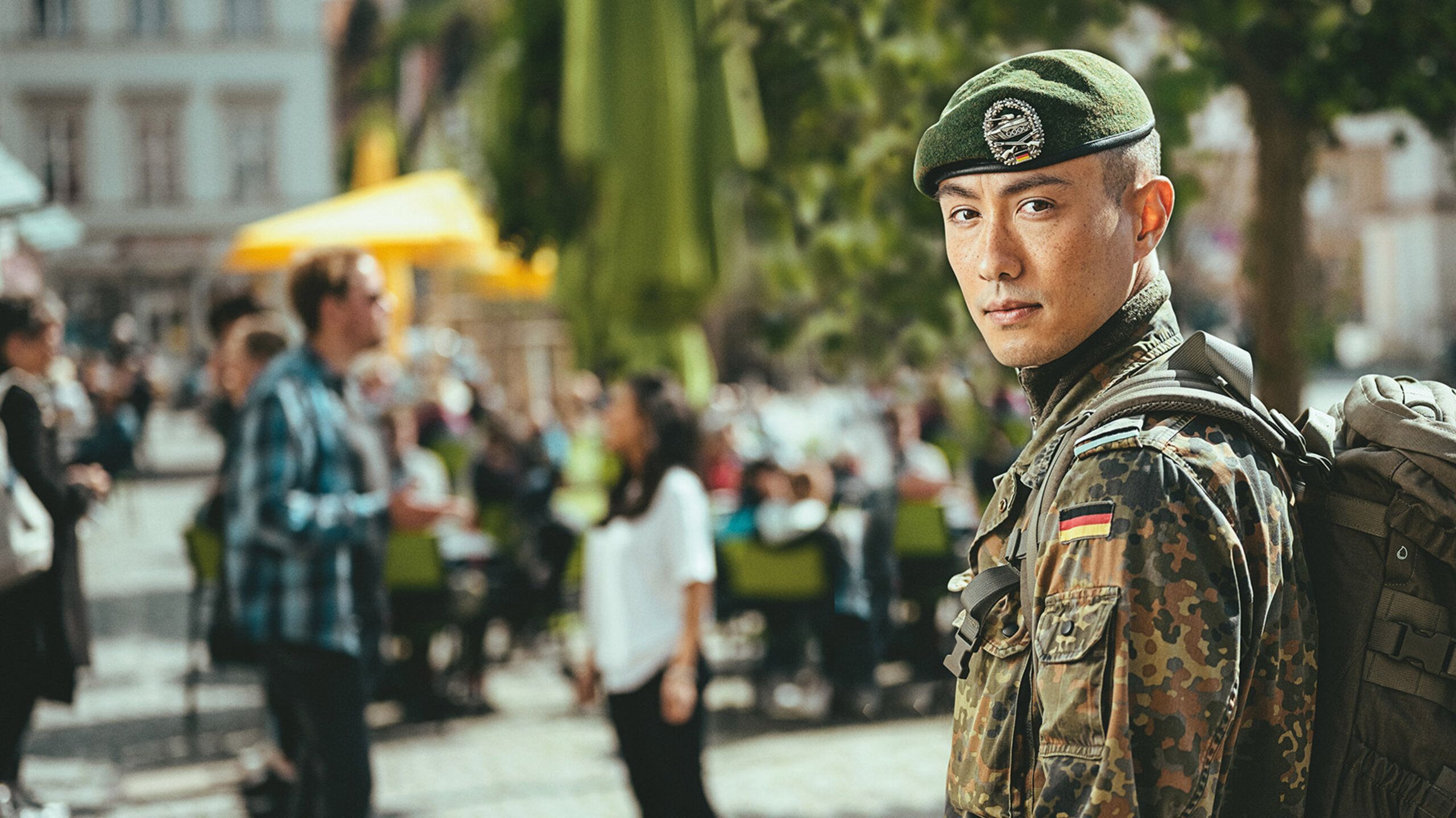 Ein Soldat in Uniform schaut über die Schulter in die Kamera, im Hintergrund sind Menschen in einer Stadt zu erkennen.