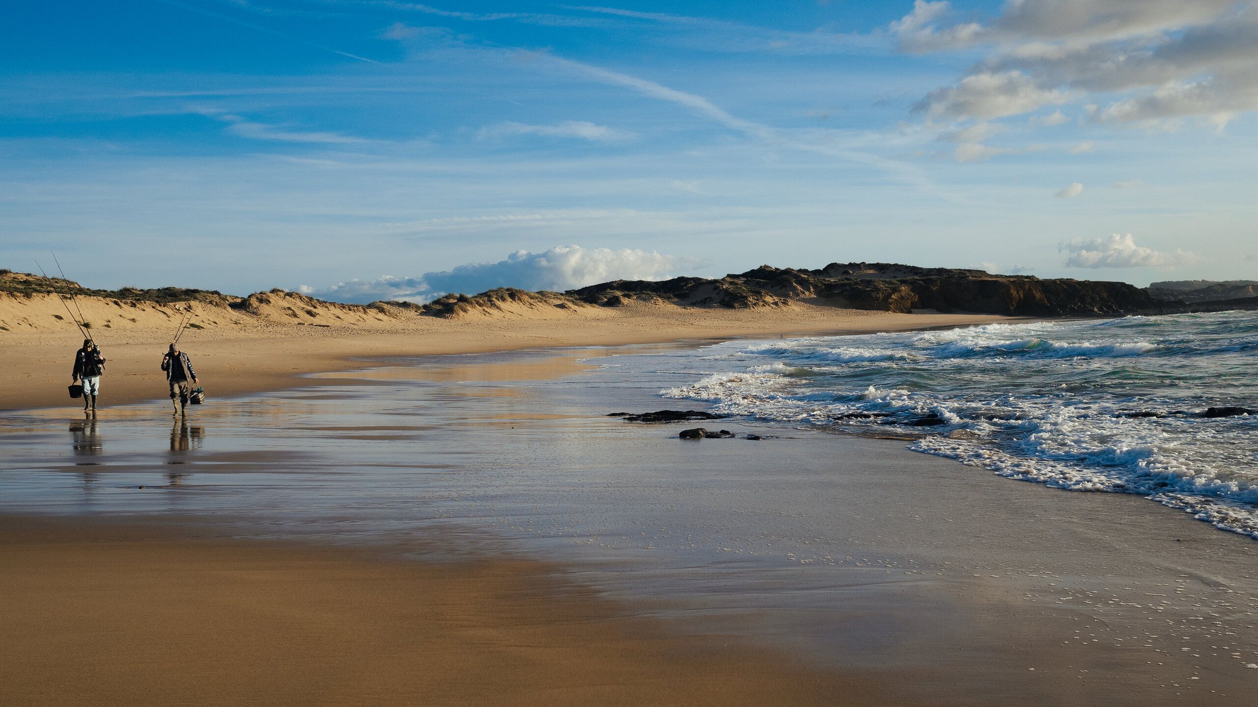 Ein Strand, an dem zwei Menschen stehen. Rechts die Wellen, im Hintergrund ein Hügelkette