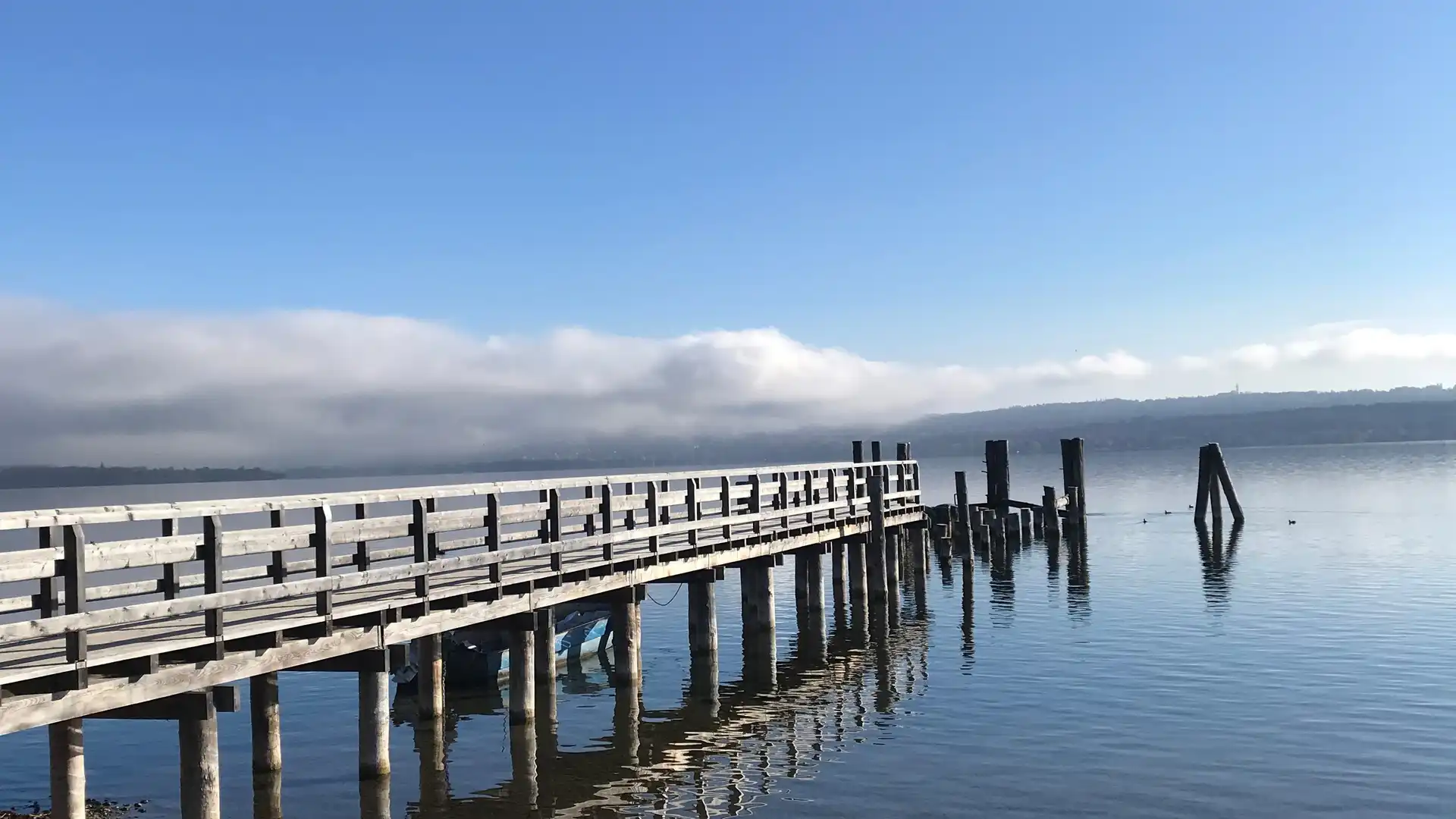 Ein Holzsteg der in das Wasser eines Sees reicht. Im Hintergrund sind Berg unter Wolke zu erkennen.