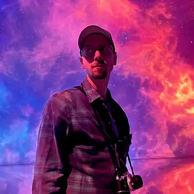 Adrian Schmidt, Mitarbeiter der Castenow GmbH, trägt eine Fotokamera um den Hals und eine Kappe auf dem Kopf. Im Hintergrund sind Lichteffekte, die einer Explosion ähneln.