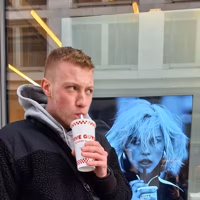 Simon Steffens, ein Mitarbeiter der Castenow GmbH, steht vor einem Schaufenster und trinkt aus einem Becher mit Strohhalm.